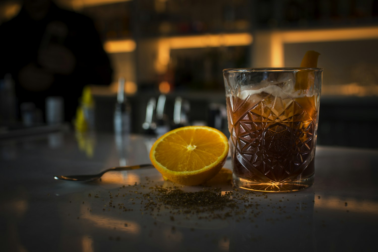 Bicchiere intagliato con drink ambrato, mezza arancia e cannella sul bancone in penombra